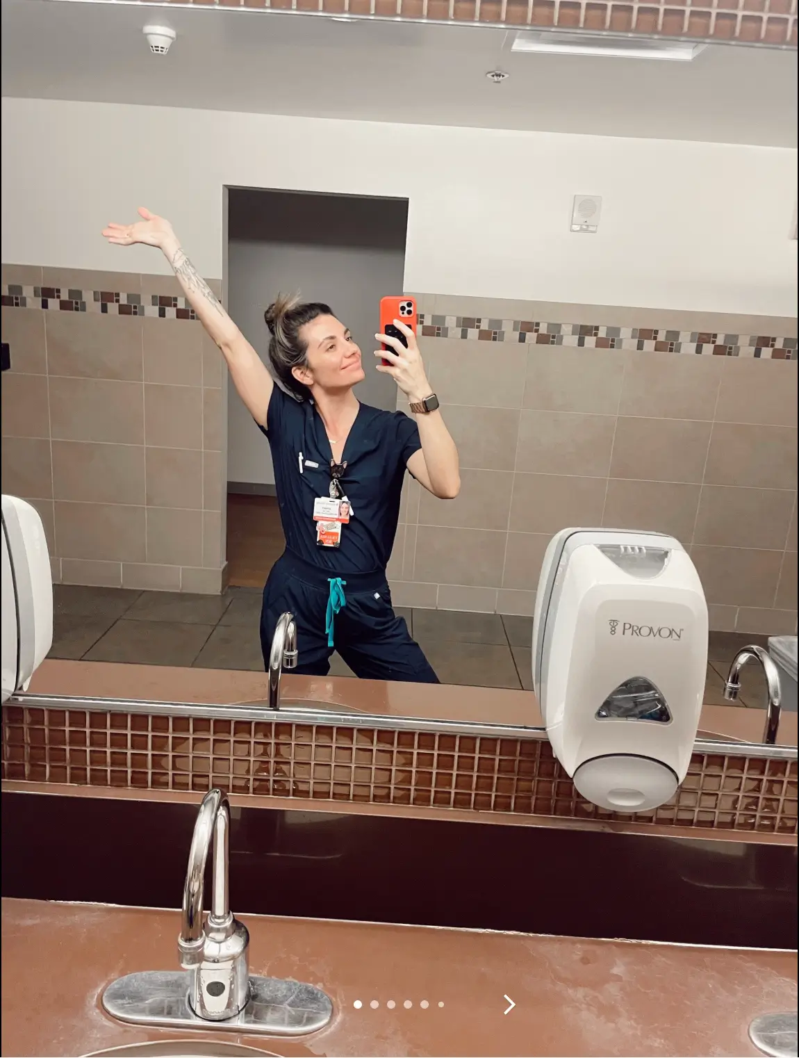 A person in medical scrubs poses cheerfully for a mirror selfie in a restroom, raising one arm and smiling. Soap dispensers and a sink are visible in the foreground.